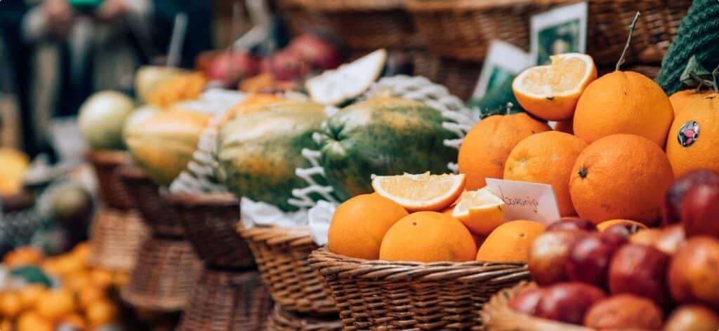Baskets of assorted fruits, including oranges, melons, and apples, are displayed on a market stall. Some oranges are sliced open, surrounded by a bustling market atmosphere. A small sign reads "Juicy."