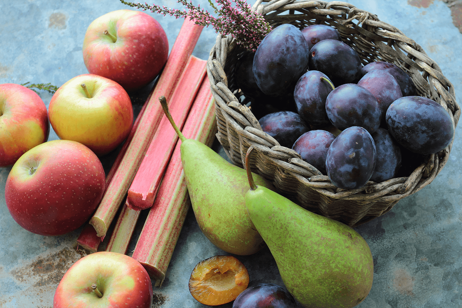 Seasonal apples, rhubarb, pears and plums in a small wicker basket organised on a dark table.