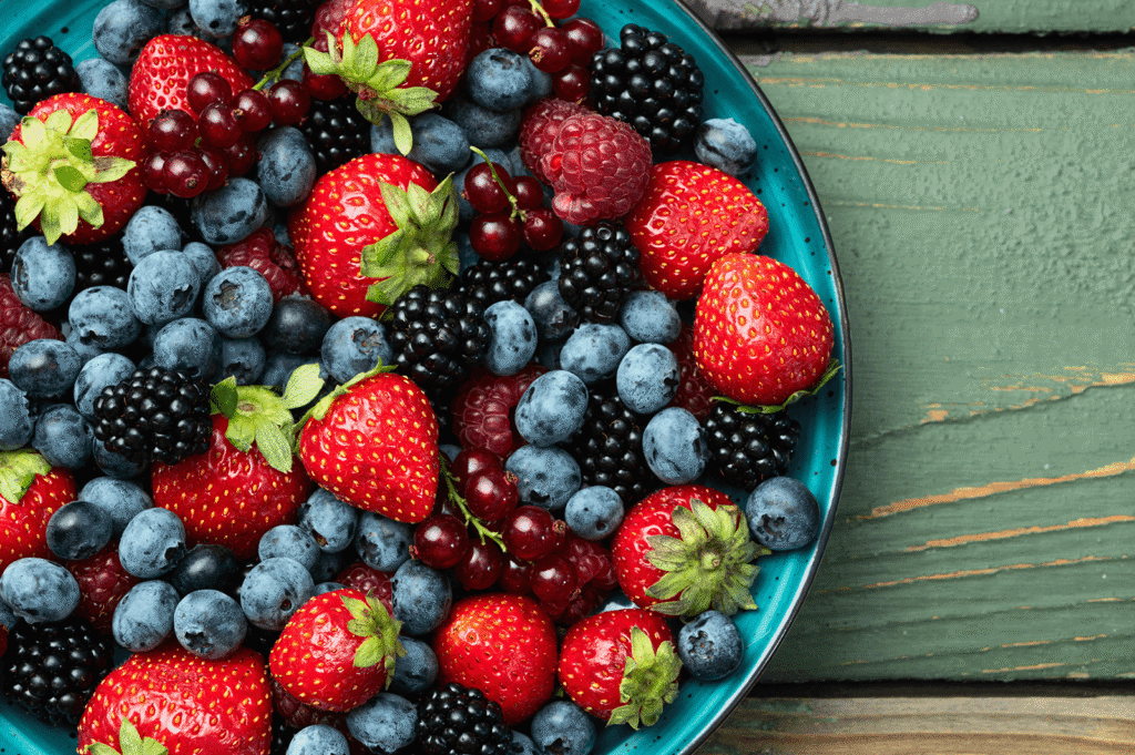 Vibrant fresh strawberries, blueberries and blackberries in a blue ceramic bowl on a green wooden table.
