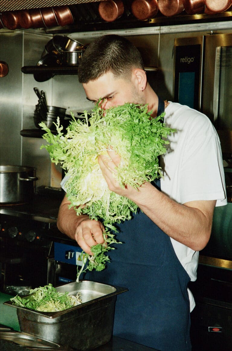A chef holds a head of frisée lettuce, tearing leaves into a metal container, in a professional kitchen with copper pans and cookware on shelves.