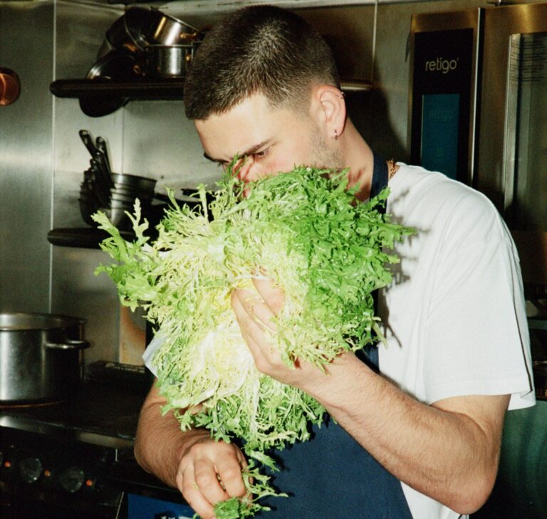 A chef holds a head of frisée lettuce, tearing leaves into a metal container, in a professional kitchen with copper pans and cookware on shelves.